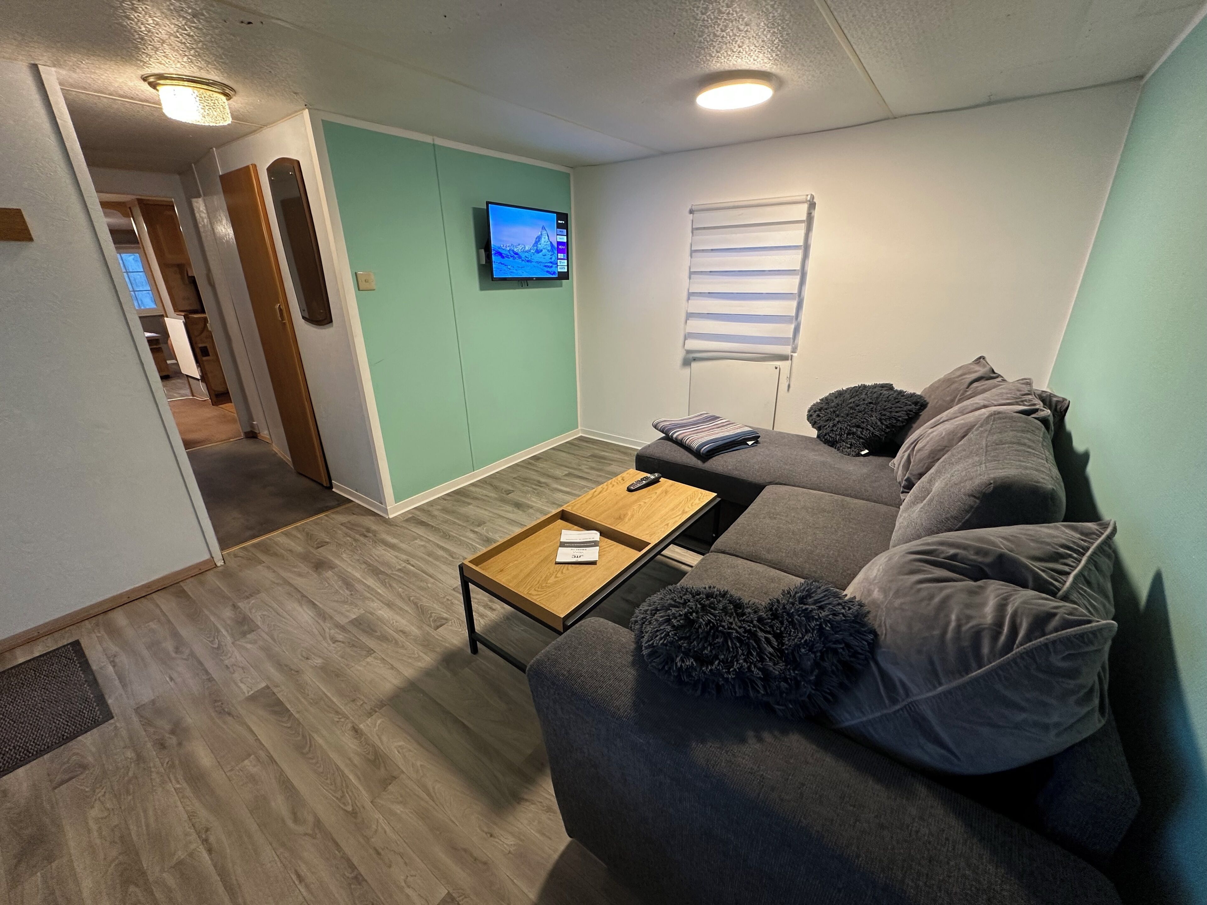 Een woonkamer met een hoekbank, een salontafel, een flatscreen televisie aan de muur en een raam met jaloezieën.