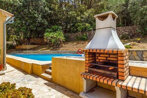 A barbecue grill with a chimney, a swimming pool, and a stone wall in the background. - Villa "Alexandria" with Mountain View, Pool, A/C, Terrace & Garden (Cala Sant Vicenç)