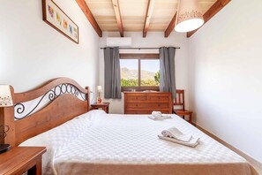 A bedroom with a wooden bed, a dresser, a chair, a nightstand, and a window with a view of the outdoors. - Villa "Alexandria" with Mountain View, Pool, A/C, Terrace & Garden (Cala Sant Vicenç)