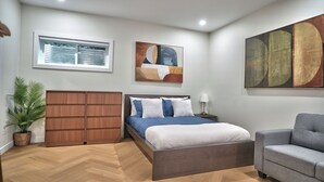 Ein Schlafzimmer mit einem Bett, einem grauen Sessel, einem Holzschrank, einer Topfpflanze und abstrakter Wandkunst.