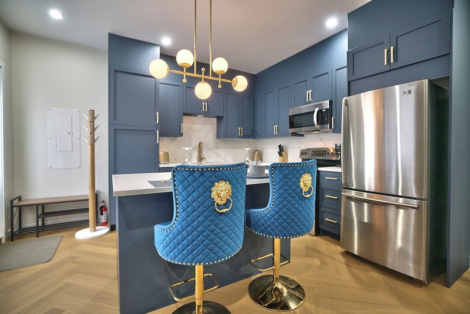 A modern kitchen with dark blue cabinets, a stainless steel refrigerator, a double sink, and two blue bar stools with gold accents.