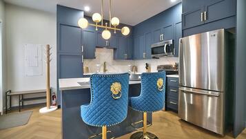 A modern kitchen with dark blue cabinets, a stainless steel refrigerator, a double sink, and two blue bar stools with gold accents.