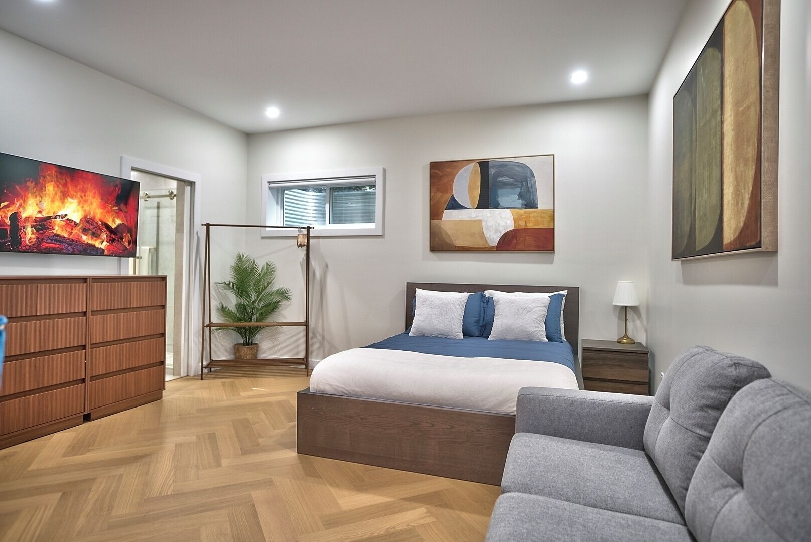 A modern bedroom with a bed, a grey sofa, a wooden dresser, a potted plant, and artwork on the walls.