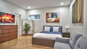 A modern bedroom with a bed, a grey sofa, a wooden dresser, a potted plant, and artwork on the walls.