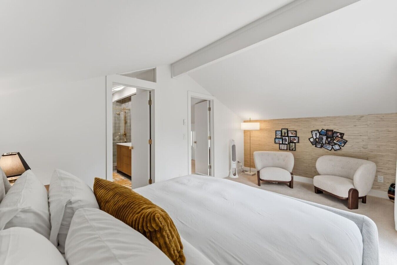 A bedroom with a bed, two chairs, a wall-mounted picture collection, and a wooden accent wall.
