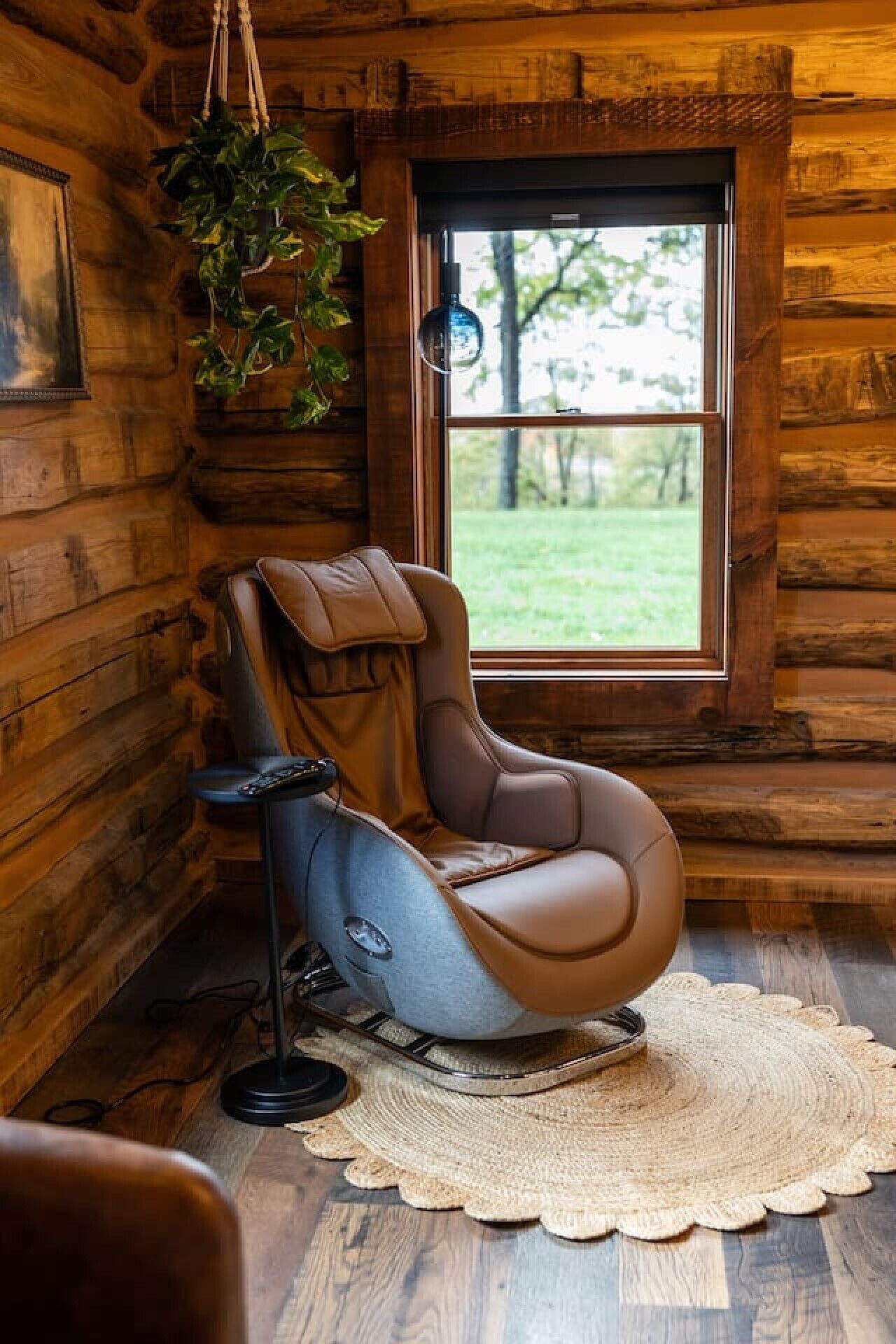 Een massagestoel in een blokhutkamer met een raam, een plant en een vloermat.