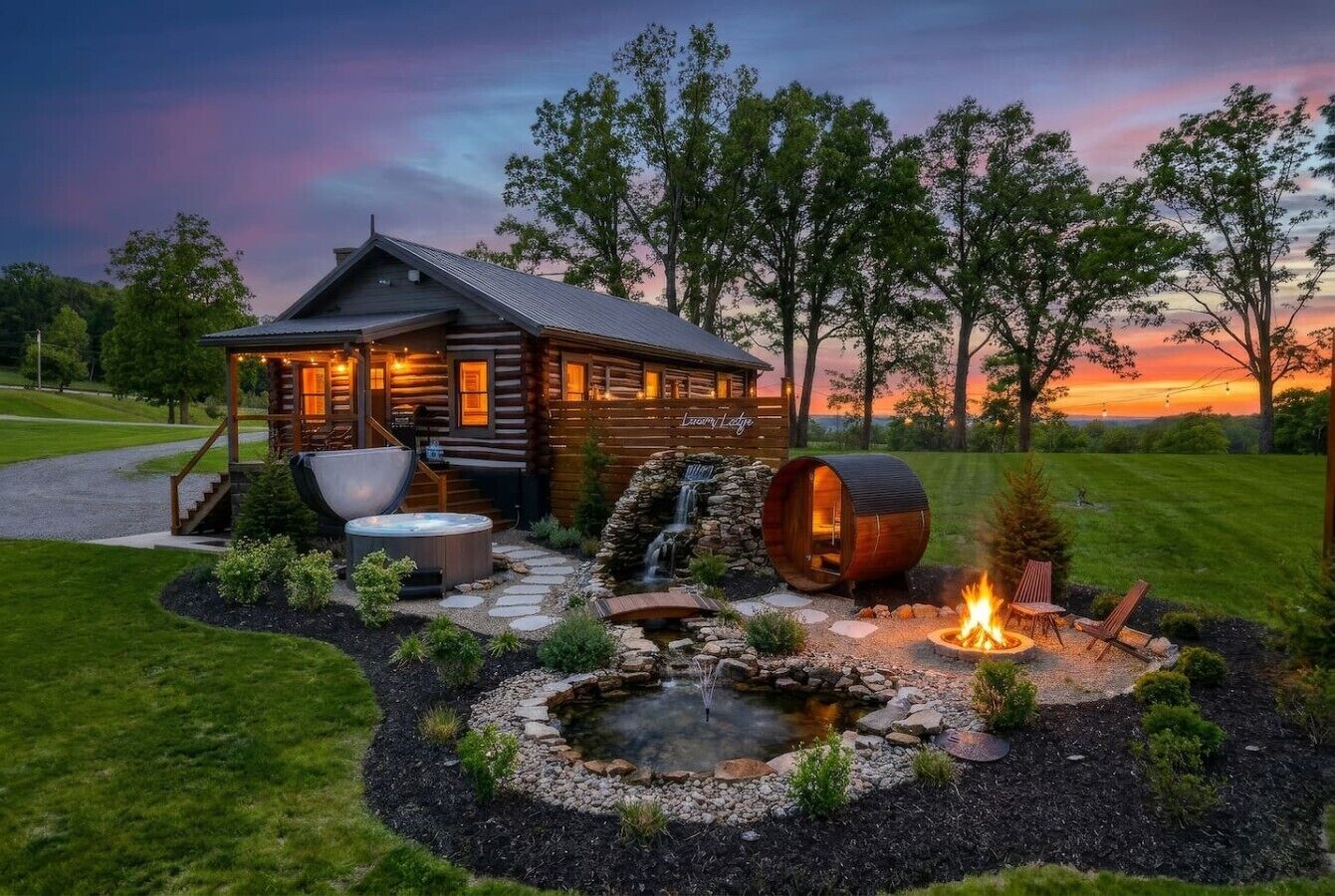 Una cabaña de madera con una cascada de piedra, un jacuzzi y una fogata rodeada de un jardín ajardinado.