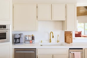 Eine Küche mit weißen Schränken, einem Edelstahlbackofen, einer Kaffeemaschine, einem Waschbecken mit goldenen Armaturen und einer Holz-Schneidebrett.