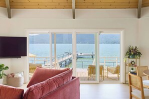Een kamer met een grote schuifpui die uitkomt op een balkon met uitzicht op een waterpartij en een steiger. Er is een televisie aan de muur bevestigd, een bank met kussens en een kleine tafel met stoelen.