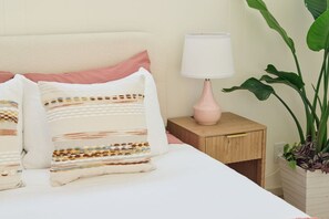 Een net opgemaakt bed met gestreepte kussens, een bedlampje en een potplant.