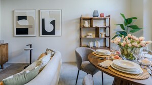 Uma área de refeições com uma mesa redonda posta para quatro pessoas, um sofá, uma estante de livros e dois quadros abstratos na parede.