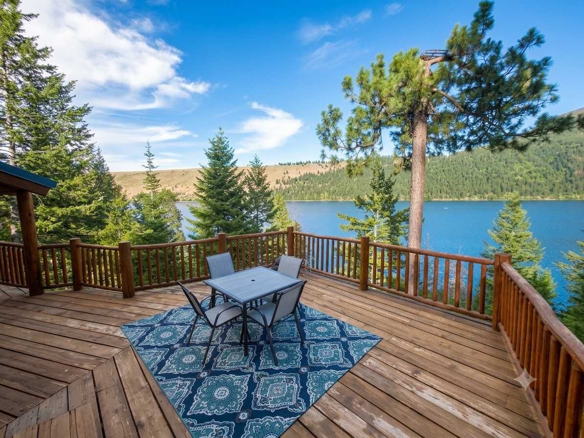 A wooden deck with a table and chairs, overlooking a lake with trees and hills in the background.