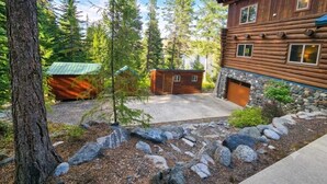 Une cabane en rondins avec une cheminée en pierre, un petit abri de jardin et un jardin bien entretenu agrémenté de rochers et d’arbres.