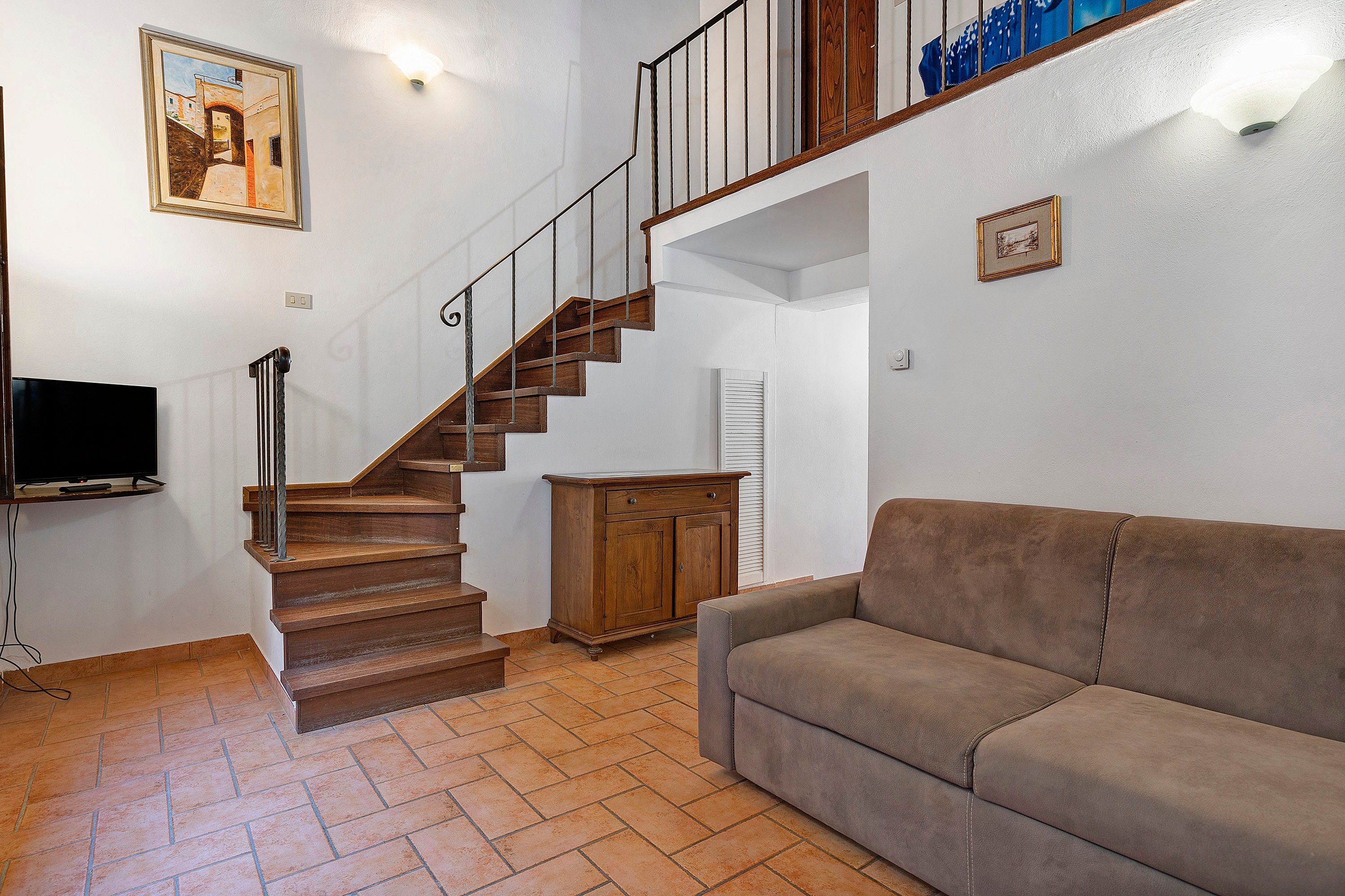 A staircase with wooden steps and metal railings, a flat-screen TV on a stand, a brown leather sofa, and a wooden cabinet.