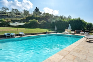 Ein rechteckiger Swimmingpool mit klarem, blauem Wasser, umgeben von einer Steindeck. Am Poolufer stehen Liegestühle und Sonnenschirme.