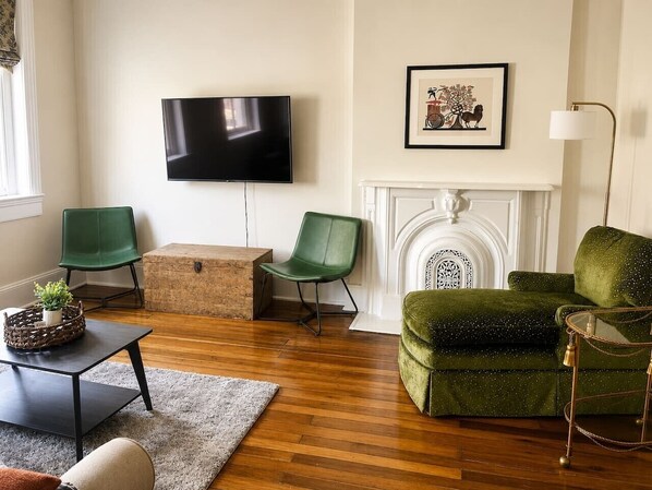 A living room with a flat-screen TV, green chairs, a green sofa, a fireplace, and a wooden floor.