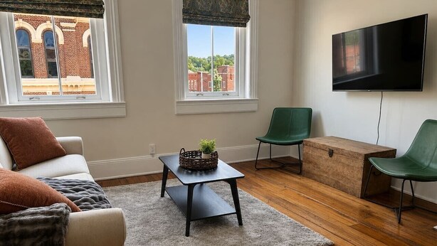 A living room with a sofa, a coffee table, and two chairs. There is a television mounted on the wall, and windows with blinds.