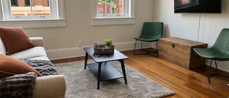 Un salon comprenant un canapĂ©, une table basse et deux fauteuils. Un tĂ©lĂ©viseur est fixĂ© au mur, et les fenĂȘtres sont Ă©quipĂ©es de stores.