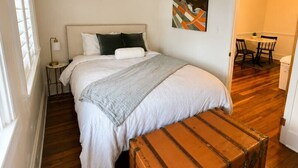 A bedroom with a bed, a bedside table, a lamp, a painting, a suitcase, and a dining area in the background.