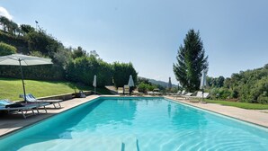 Ein rechteckiger Pool mit klarem, blauem Wasser, umgeben von Liegestühlen und Sonnenschirmen, vor einer Kulisse aus Bäumen und einer Steinmauer.