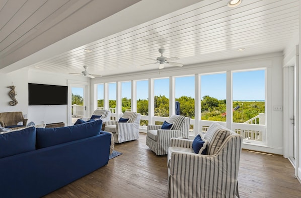 A spacious room with a large window, a ceiling fan, and a blue sofa. - Stunning Oceanfront Home, Guest Cards & Golf Cart (Fripp Island)