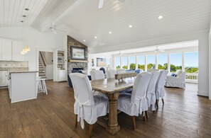 Un vaste espace de salle à manger avec une table en bois et des chaises blanches, une cuisine dotée de meubles blancs et une vue sur l’extérieur grâce à de grandes fenêtres.