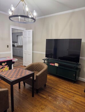 A living room with a wooden floor, a television on a stand, a chair, and a table with a chessboard. - Magnificent Morningside Manor (Vidalia)