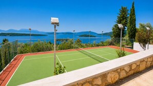 Uma quadra de tênis com muro de pedra e cerca de metal, com vista para um lago e montanhas.