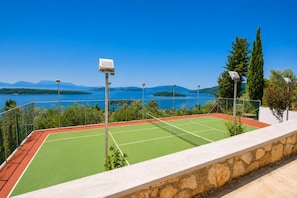 Ein Tennisplatz mit Steinmauer und Metallzaun, mit Blick auf einen See und Berge.
