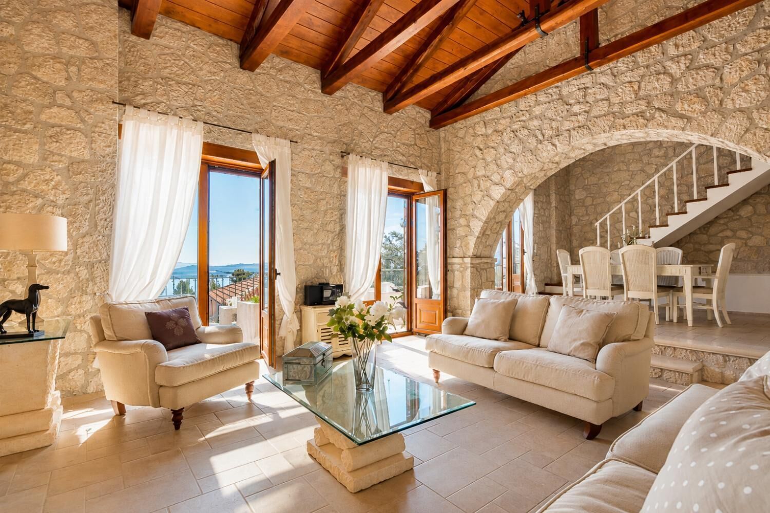 A spacious living room with stone walls, wooden beams, a glass coffee table, and a view of the outdoors.