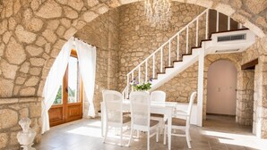 Um quarto com paredes de pedra, uma mesa de jantar, cadeiras, uma escadaria e um candelabro.