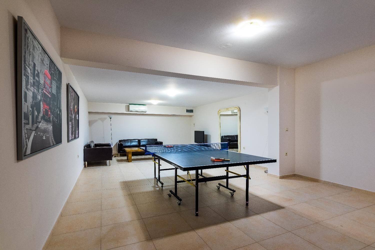 Ein Zimmer mit Tischtennisplatte, einer schwarzen Ledersofa, einem Fernseher und einem Spiegel.