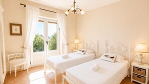 Quarto de hotel com duas camas, uma cadeira, janela com cortinas, lustre e mesa de cabeceira com abajur.