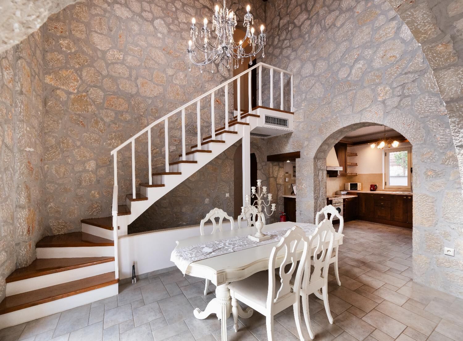 A stone-walled interior with a white staircase, a dining table with chairs, and a chandelier.
