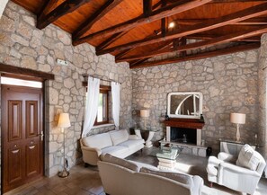 A stone-walled room with a wooden ceiling, a sofa, two armchairs, a fireplace, and a door. - Villa Galatia - Three Bedroom Villa, Sleeps 6 (Perigiali)