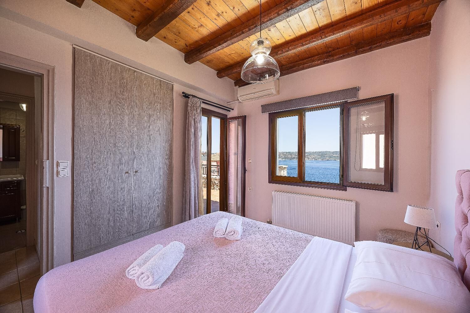 A bedroom with a large bed, a view of the sea, a wooden ceiling, and a window with a mirror.