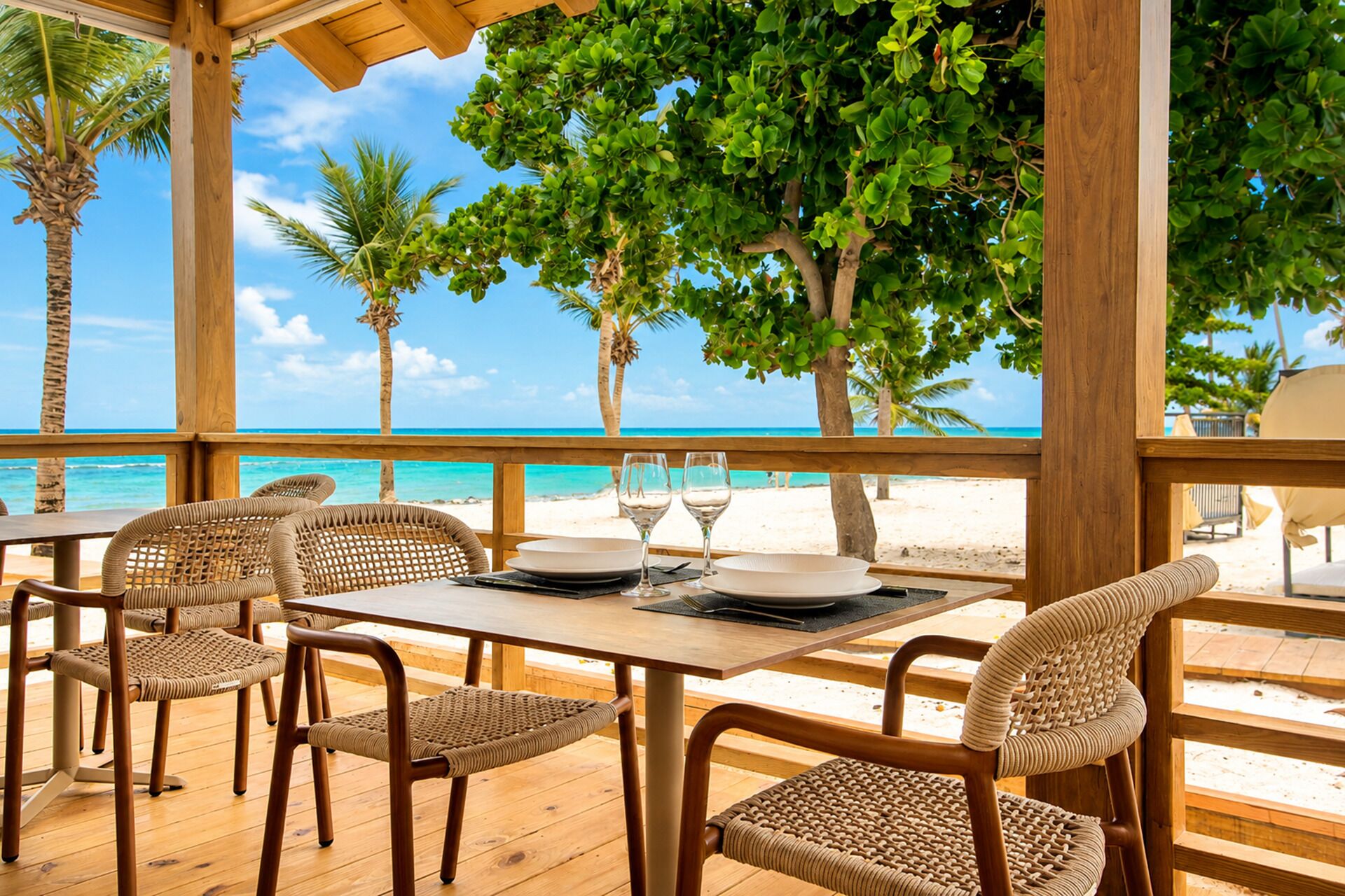 An outdoor dining area with a wooden table and wicker chairs, overlooking a beach with palm trees and the ocean.