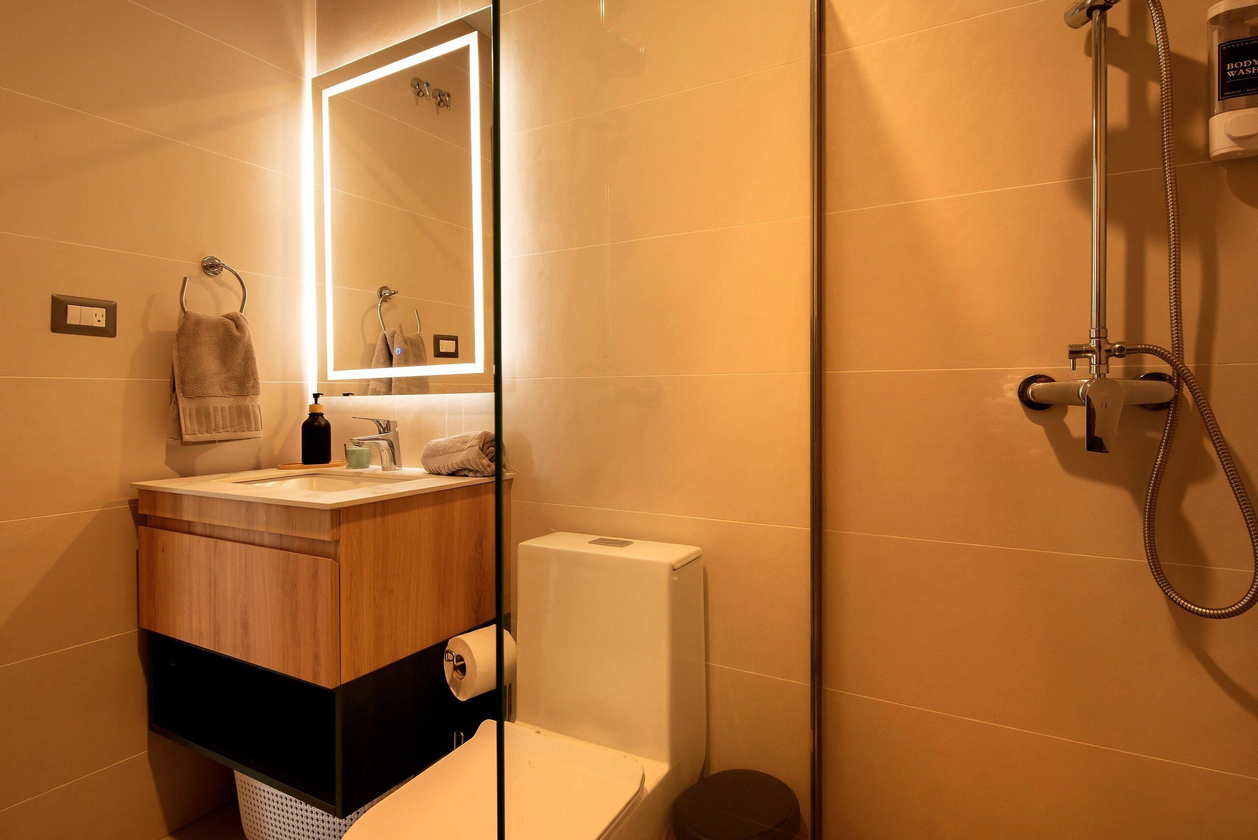 A bathroom with a wooden vanity, a toilet, a shower, and a mirror with built-in lighting.