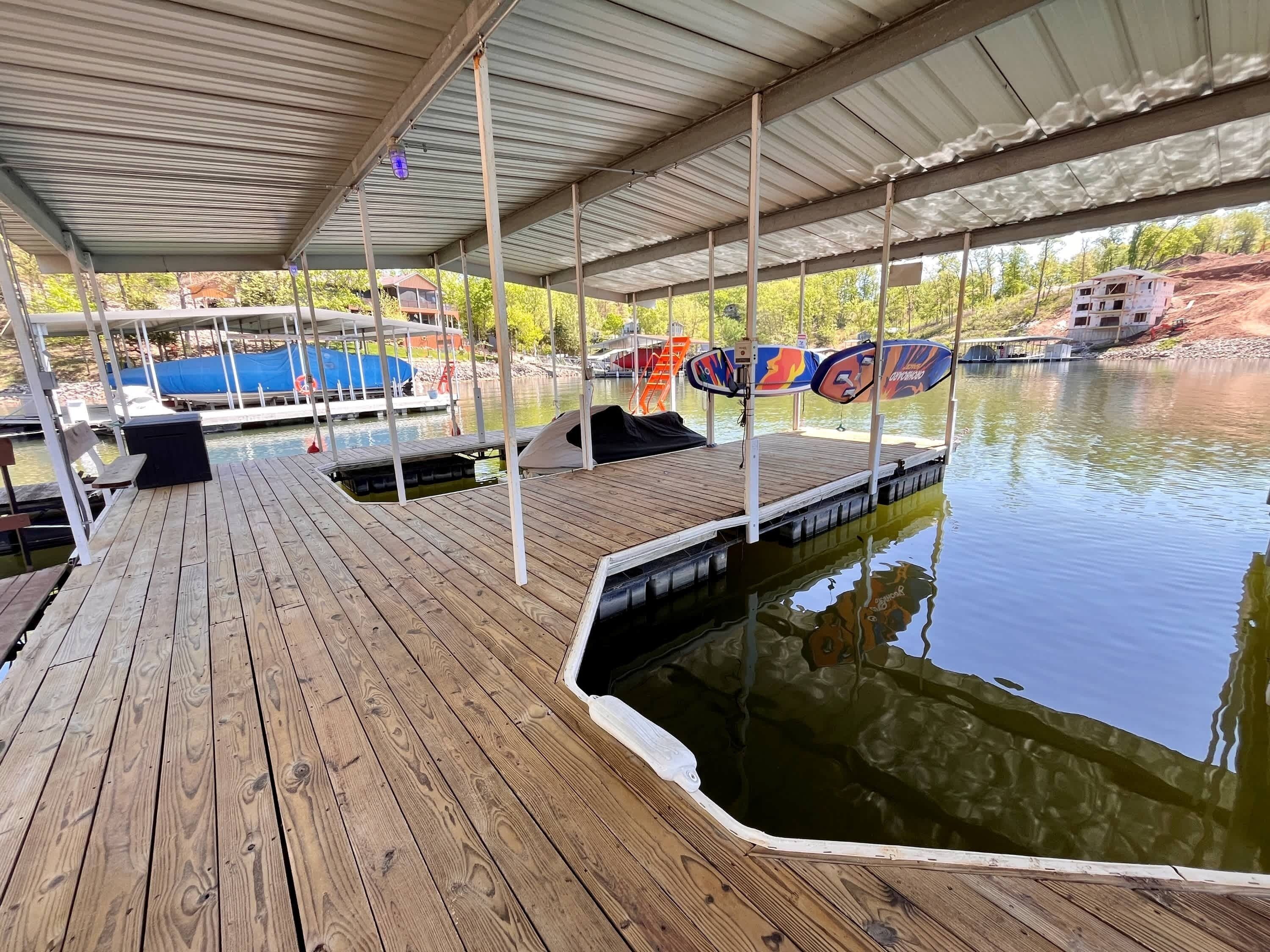 A covered wooden dock with a boat, a covered seating area, and a view of a house under construction across the water.