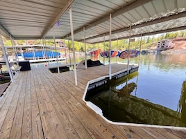 Un muelle de madera cubierto con un bote, una zona de estar techada y vistas a una casa en construcción al otro lado del agua.