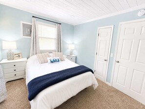 Quarto com cama, mesas de cabeceira, janela com cortinas, porta e chão coberto de carpete.