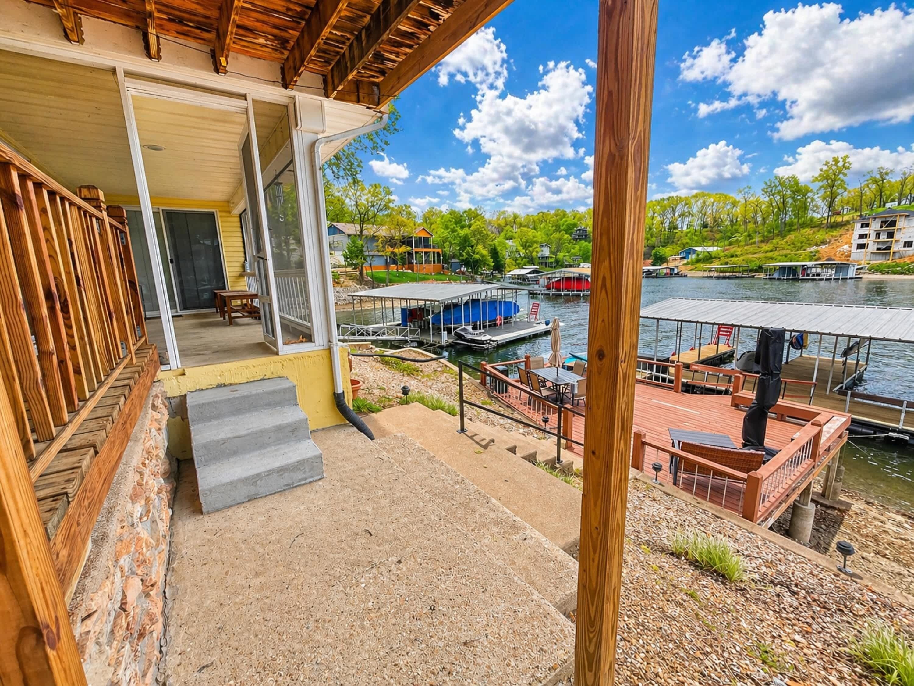 A wooden deck with steps leading to a covered porch, a view of a lake with boats, and a dock area with tables and chairs.