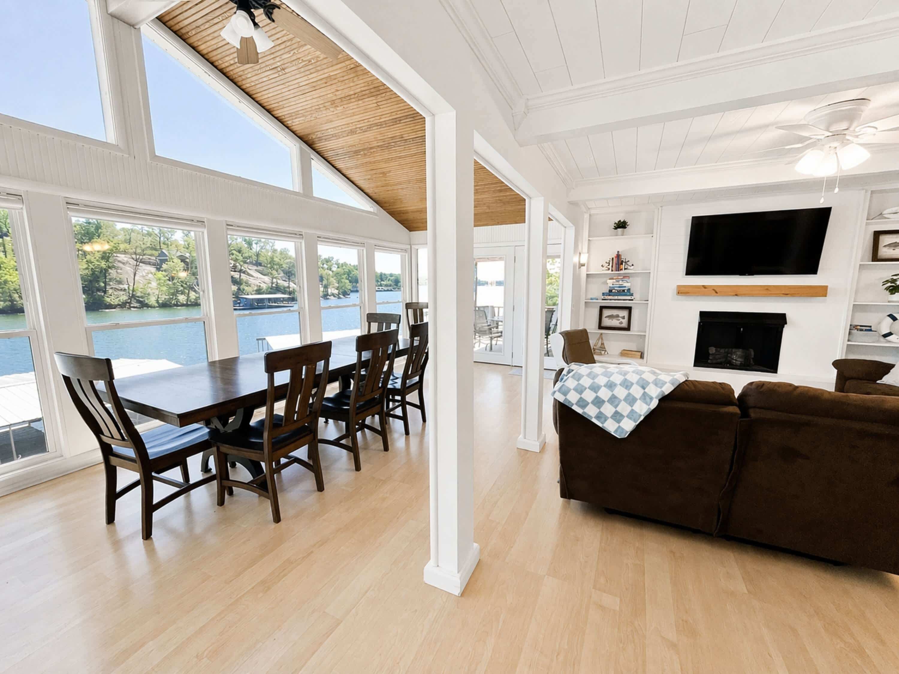A spacious living area with a dining table, brown leather couch, and a fireplace. Large windows offer a view of a lake.