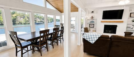 A spacious living area with a dining table, brown leather couch, and a fireplace. Large windows offer a view of a lake.