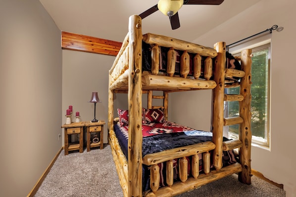 Una cama nido de madera con ventilador de techo, una mesita de noche y una ventana con vistas a árboles.