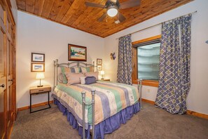 Une chambre à coucher avec un lit, un ventilateur de plafond, une fenêtre avec des rideaux et une table de chevet avec une lampe.