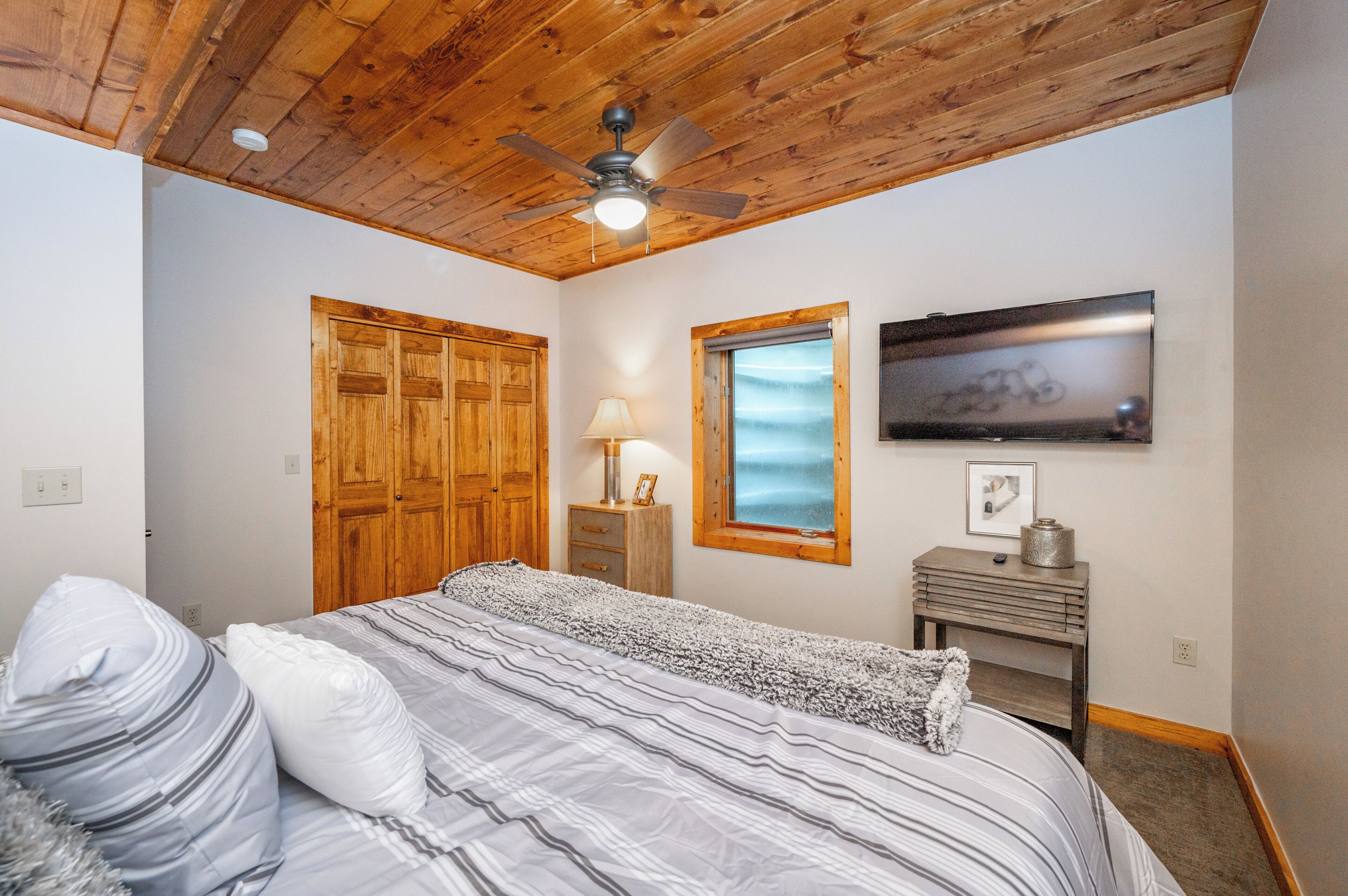 Un dormitorio con cama, armario de madera, ventilador de techo, ventana y televisor sobre un mueble de madera.