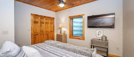 A bedroom with a bed, wooden wardrobe, ceiling fan, window, and a television on a wooden stand.