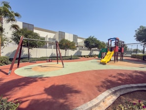 A playground with swings and a slide, surrounded by a building and trees. - Vogue, Wadi Al Safa Villa The Fields Elie Saab (Wadi Al Safa)