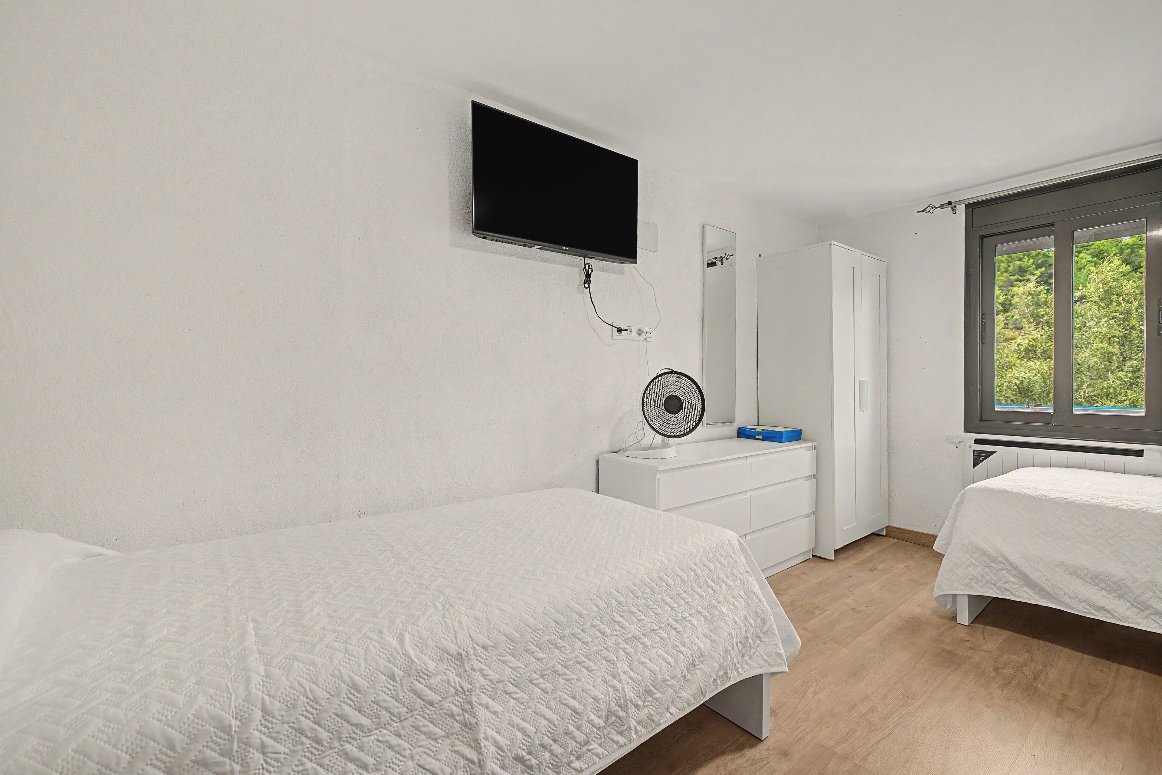 A bedroom with two beds, a television mounted on the wall, a mirror, and a window with a view of greenery.